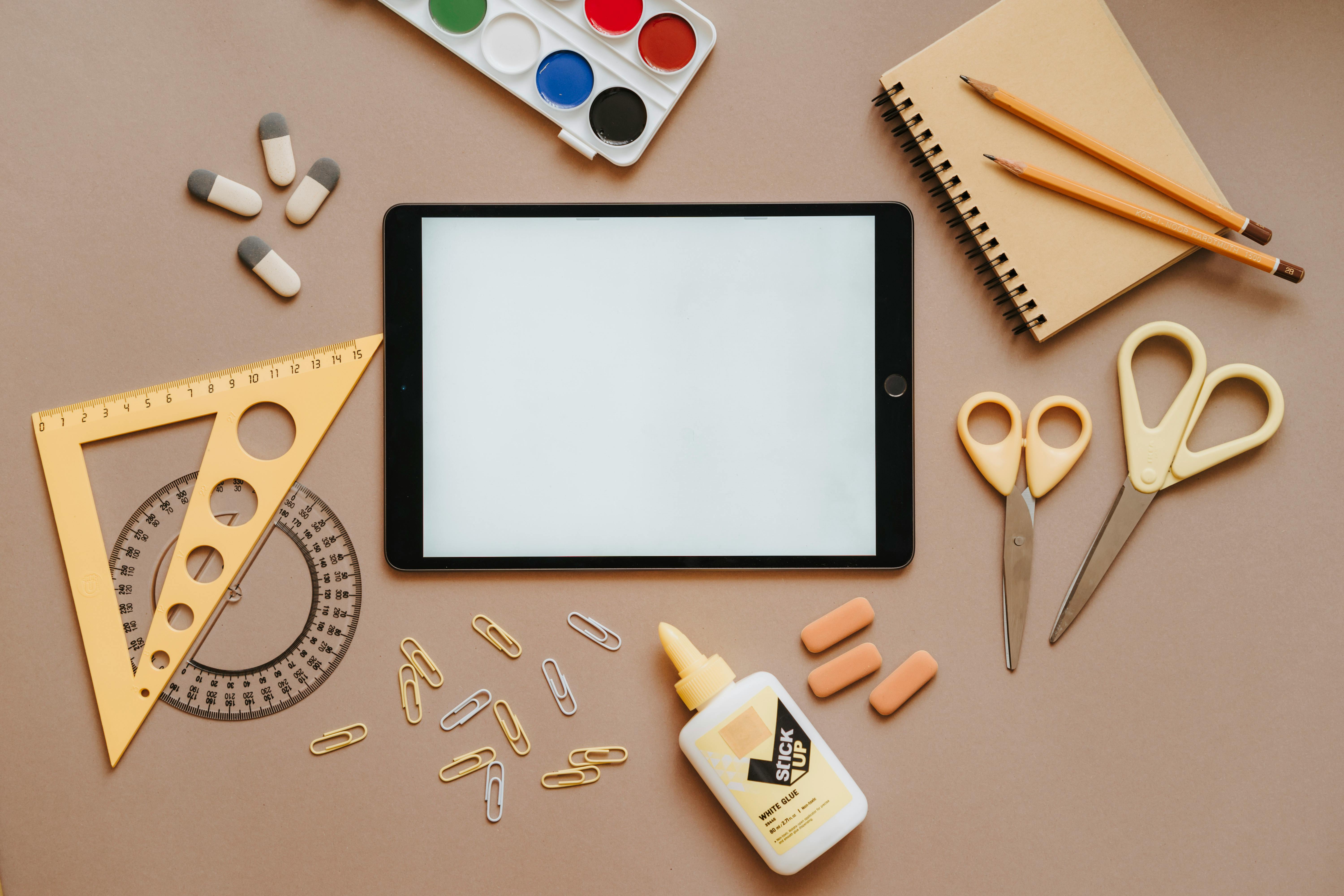 Flat lay of a tablet, notebook, glue, scissors, and paper clips arranged like a page-planning workspace